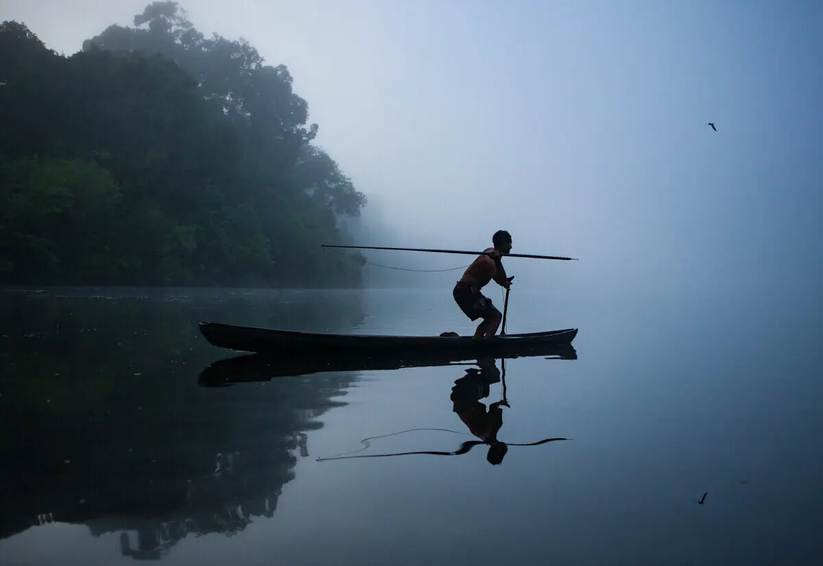 Homem em pé em uma canoa, em silhueta, sobre um rio com neblina