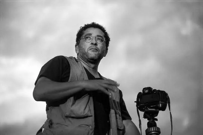 Araquém Alcântara in a black-and-white portrait, beside a camera on a tripod under a cloudy sky